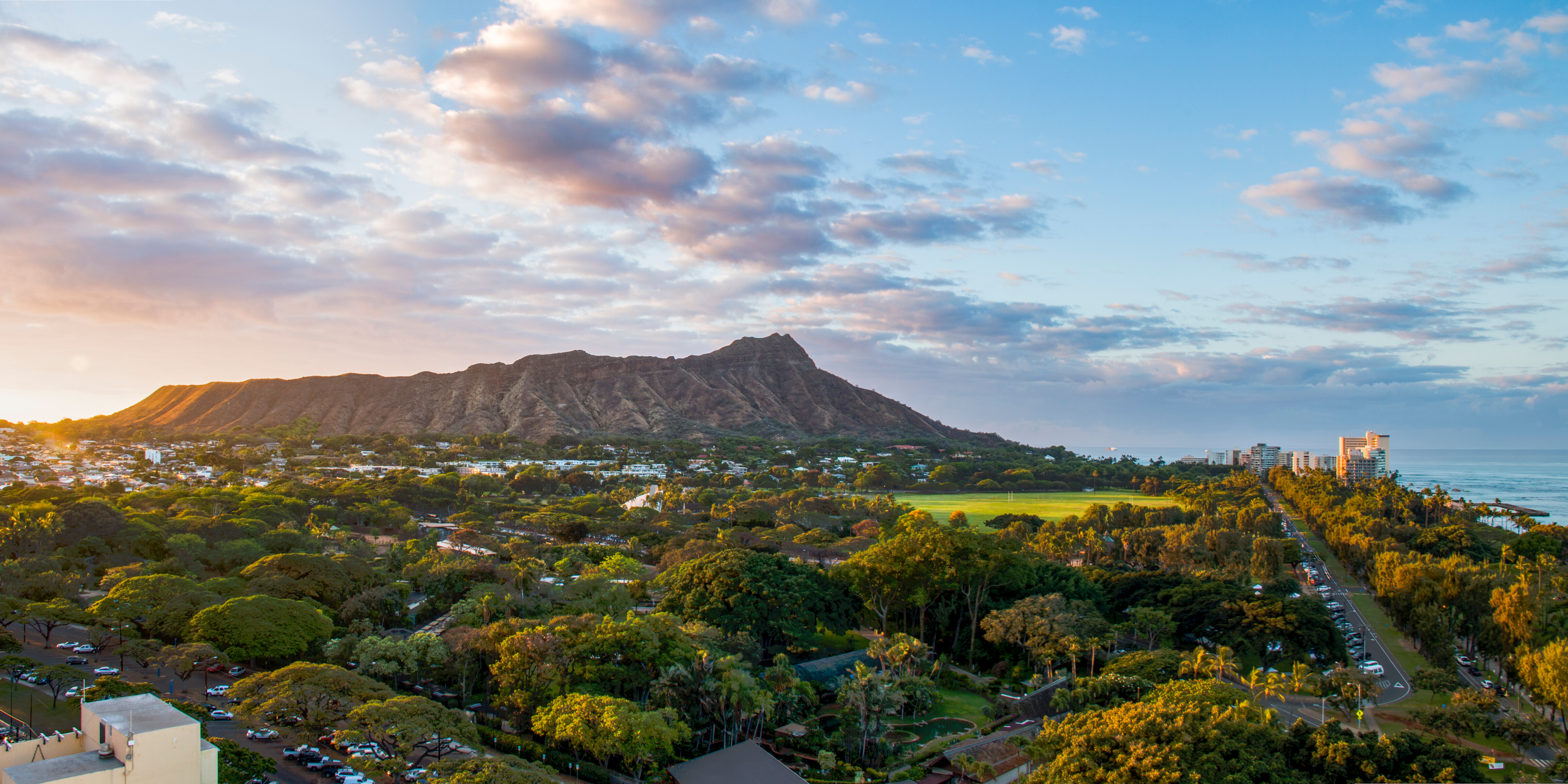 Sunlit view of a Diamond Head mountain next to a coastal cityscape with greenery and high-rises under a pastel sky.